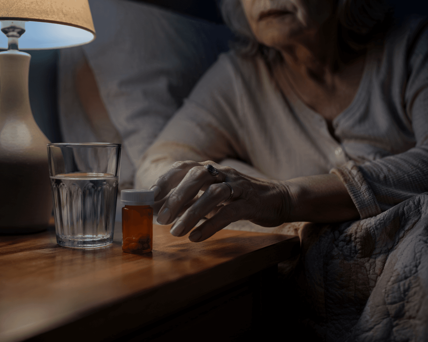 Older woman's hand near a pill bottle and glass of water on a nightstand at night