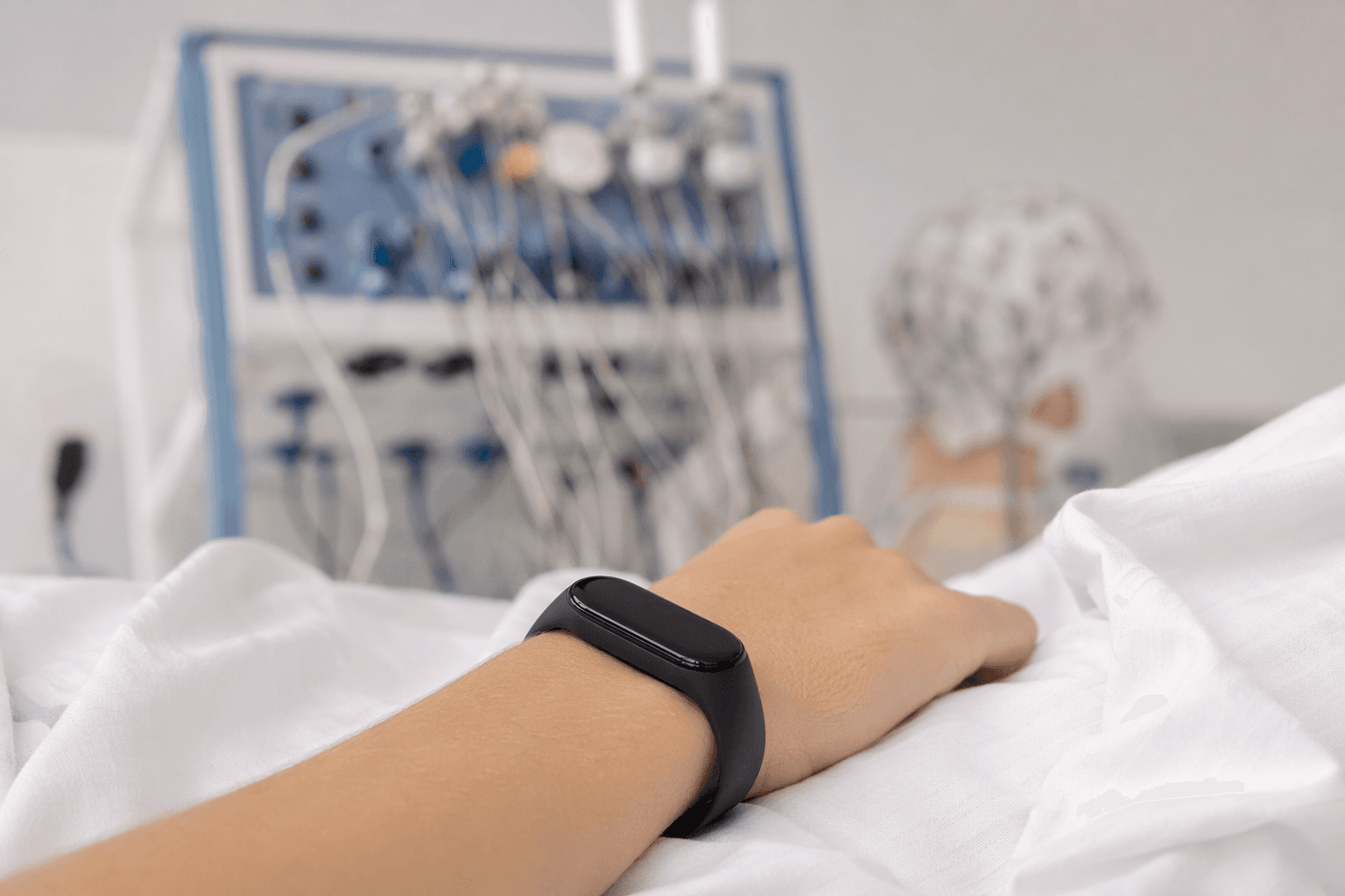 A wrist wearing a fitness tracker on a clinical bed sheet with sleep-lab equipment in the background