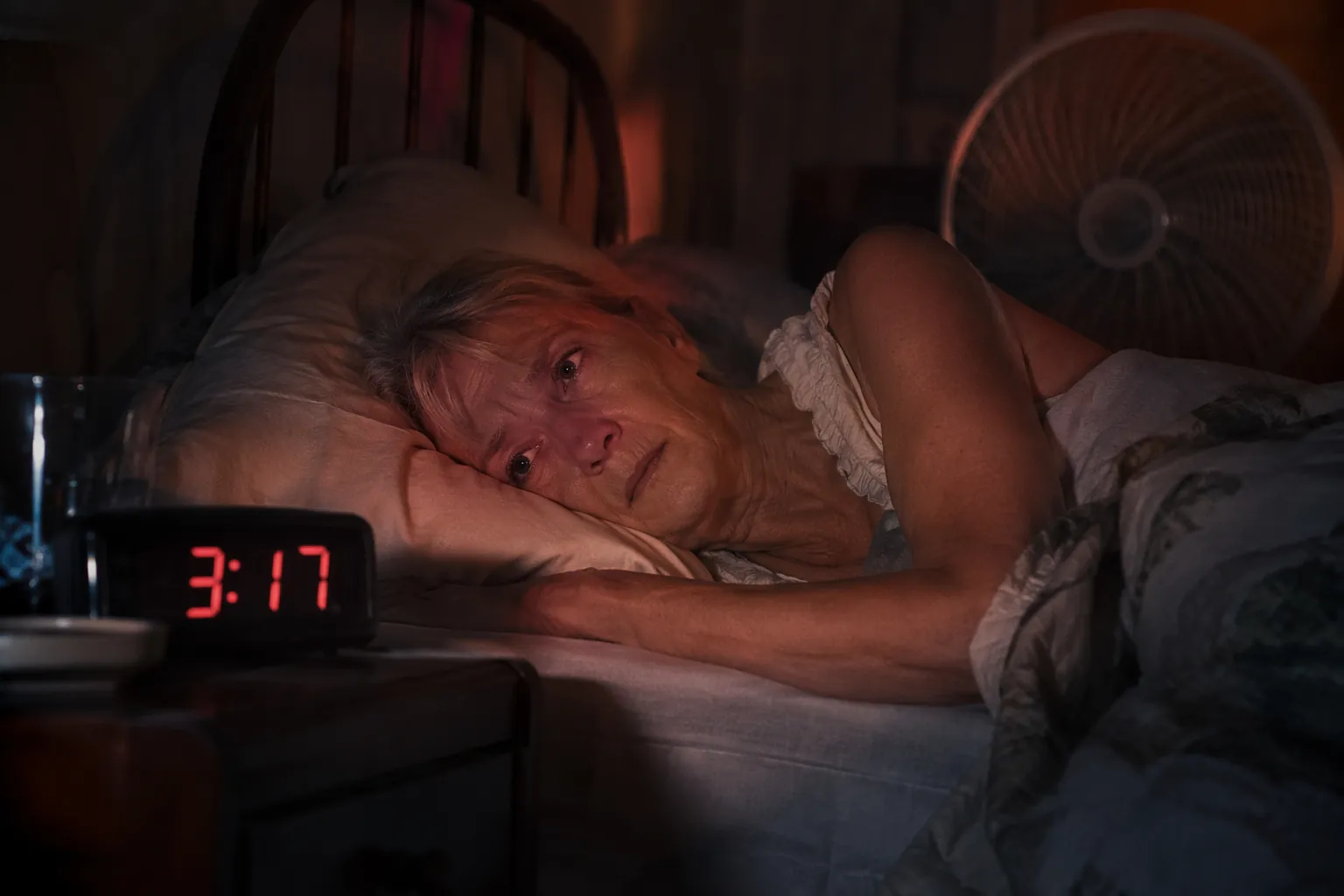 Older woman awake at night in a warm bedroom with a fan and clock showing 3:17 AM
