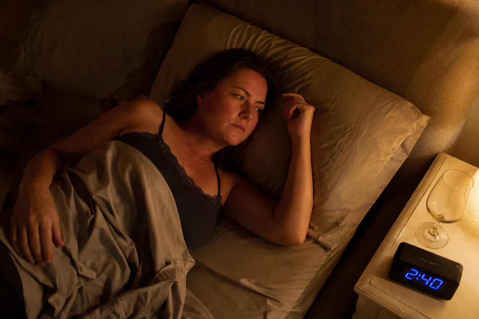 A mid-40s woman awake at 2:40 a.m. looking flushed and uncomfortable with an empty wine glass on the nightstand