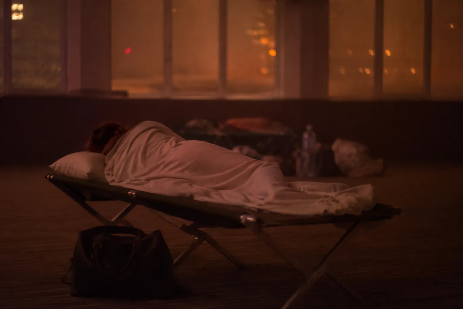A displaced person resting on a simple cot in a dim evacuation shelter