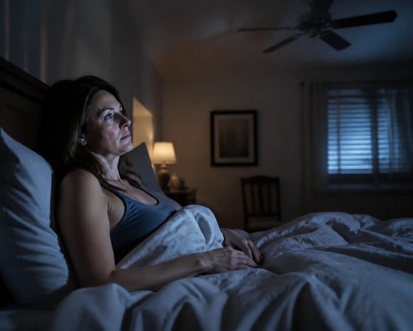 Middle-aged woman awake in bed at night with dim moonlight and a warm lamp, reflecting perimenopausal sleep disruption
