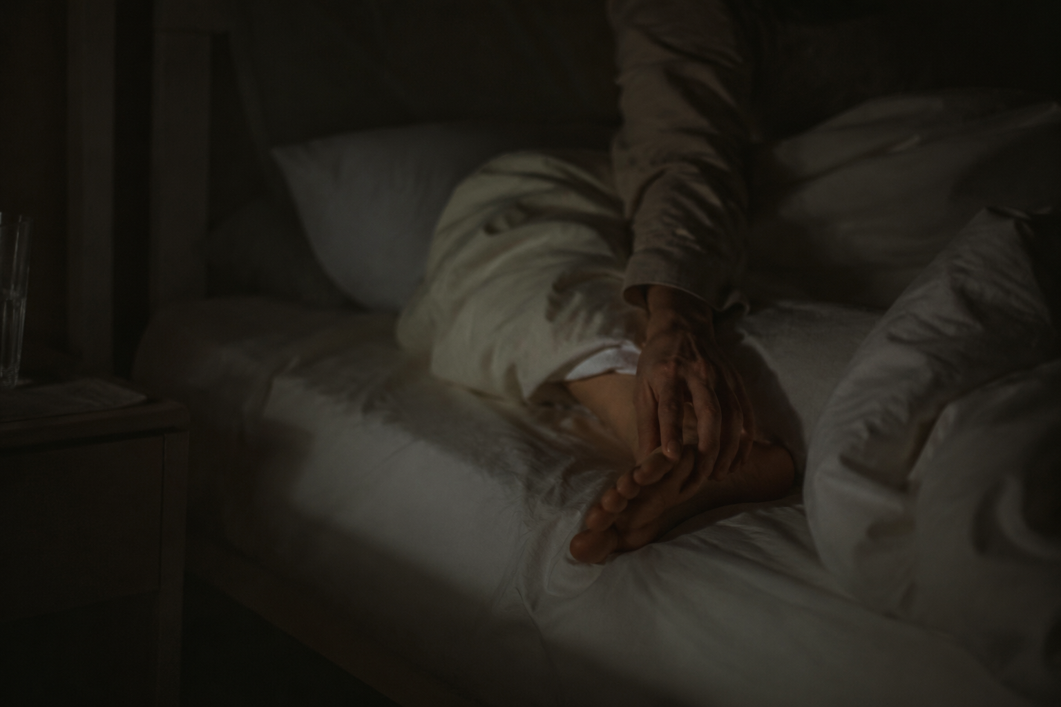 A person lying awake at night with a hand reaching toward a painful foot under the blanket