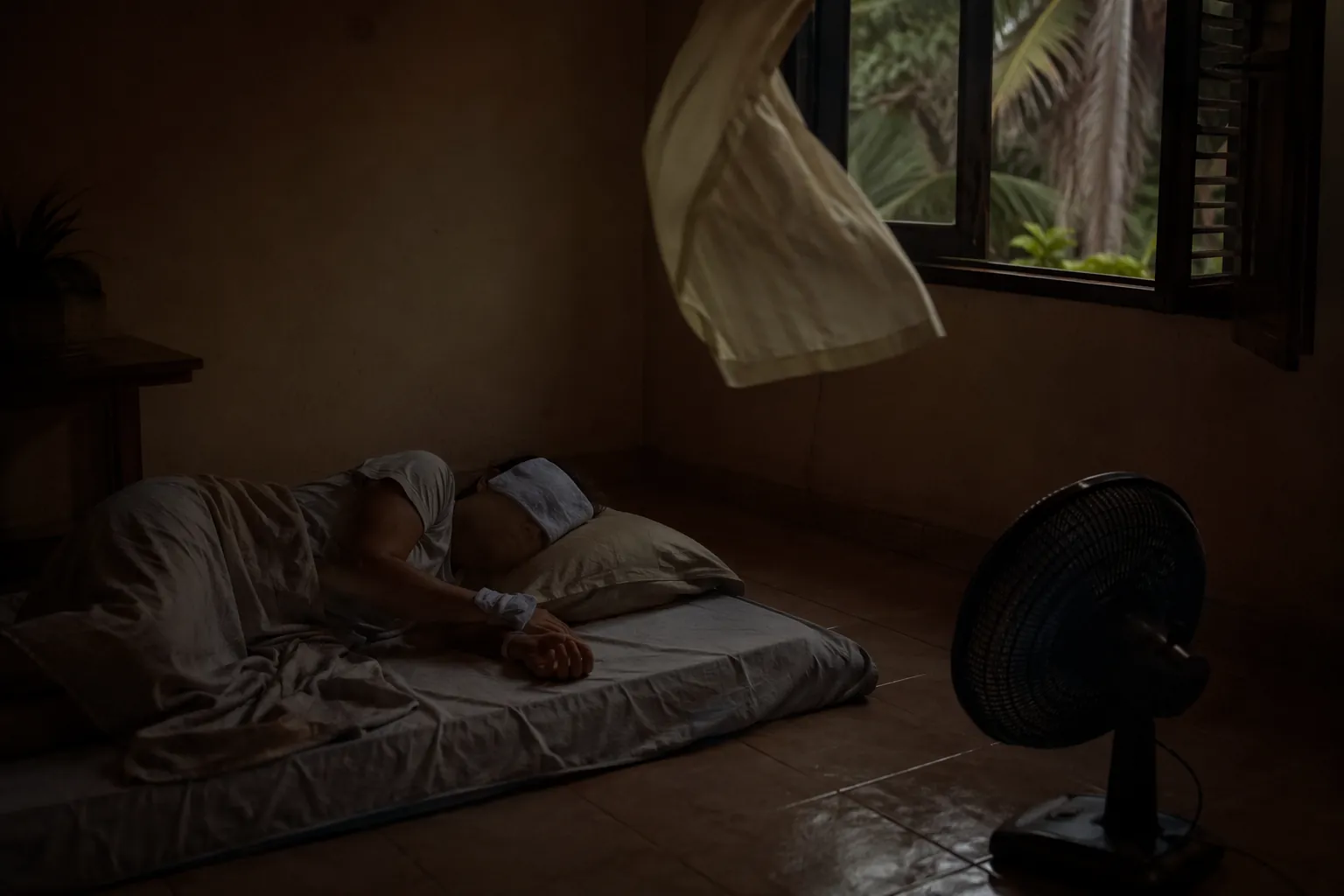 Person sleeping on a floor mattress during a power outage with a battery fan and damp cooling cloths