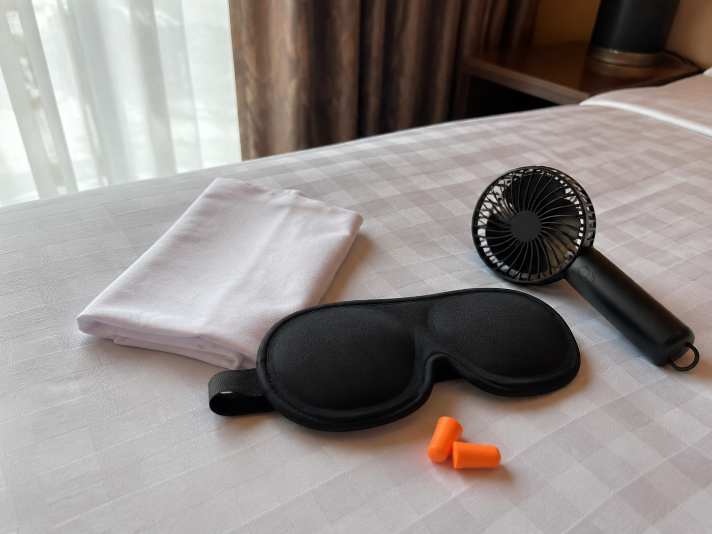 Travel sleep kit with pillowcase, contoured eye mask, earplugs, and a portable fan on a hotel bed